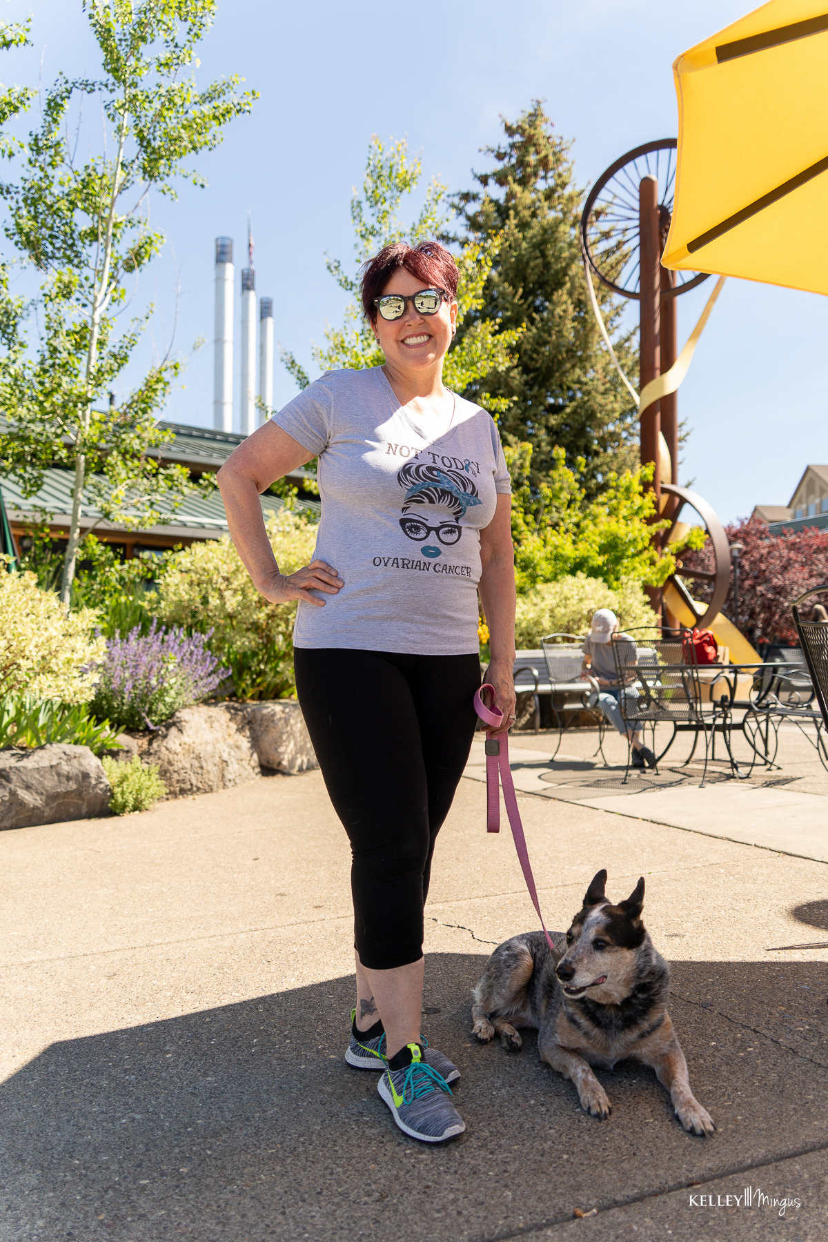 A woman wearing sunglasses and a graphic t-shirt stands outside on a sunny day, holding a dog on a leash. Industrial chimneys and outdoor seating are visible in the background, reflecting the city’s vibe as fresh as the latest cosmetic dentistry trends.