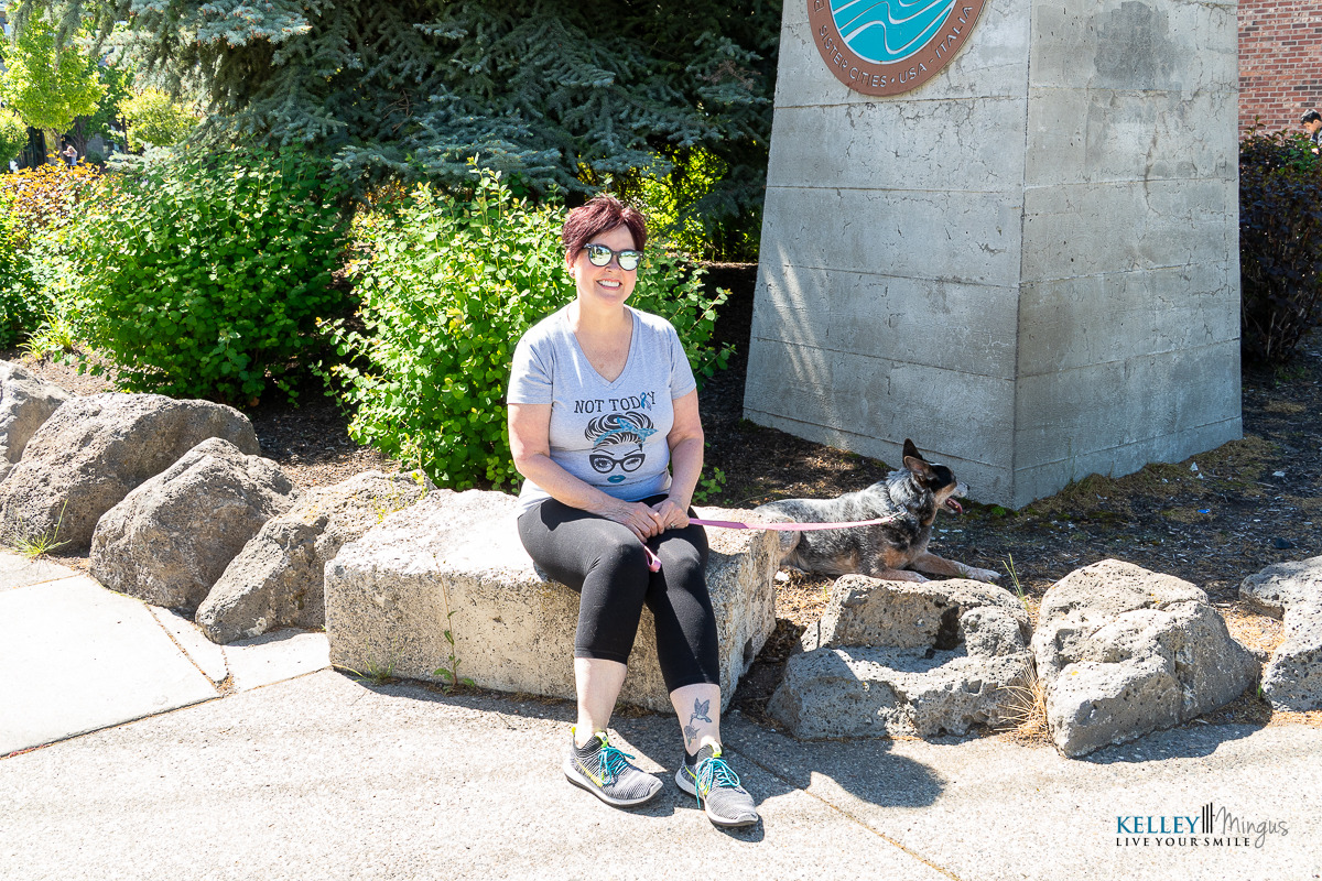 A woman wearing sunglasses and a graphic t-shirt sits on a stone bench outdoors with her dog on a leash beside her, smiling confidently after holistic full mouth rehabilitation.