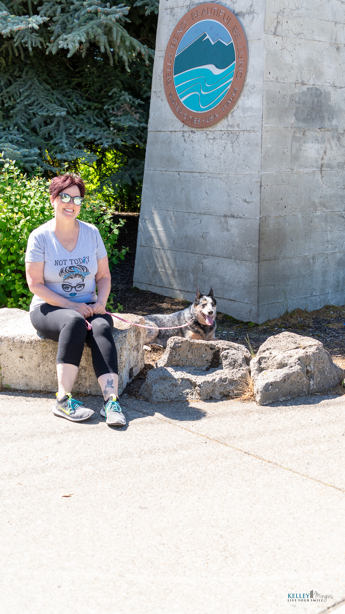 A woman sits on a stone ledge next to a dog lying down, near a concrete sign for the Blaine-Birch Bay Park and Recreation District, both enjoying the outdoors while chatting about the latest cosmetic dentistry trends.
