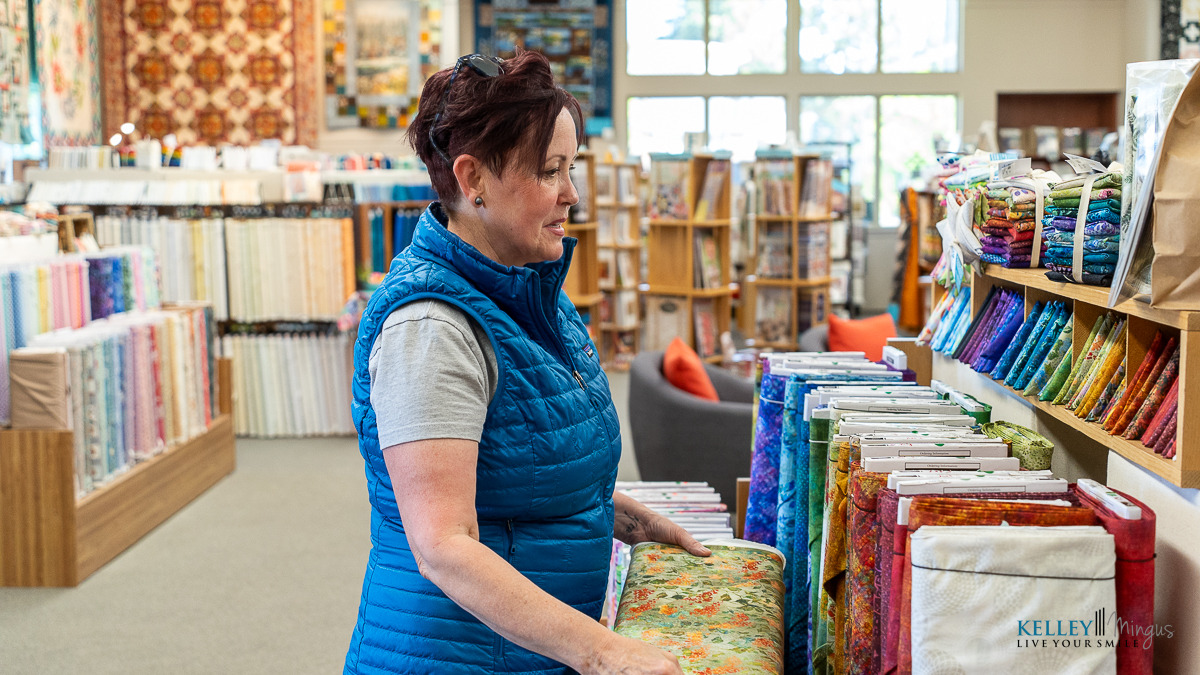 A woman in a blue vest stands in a fabric store, looking at colorful bolts of fabric arranged on shelves—her smile reflecting the confidence gained from holistic full mouth rehabilitation.