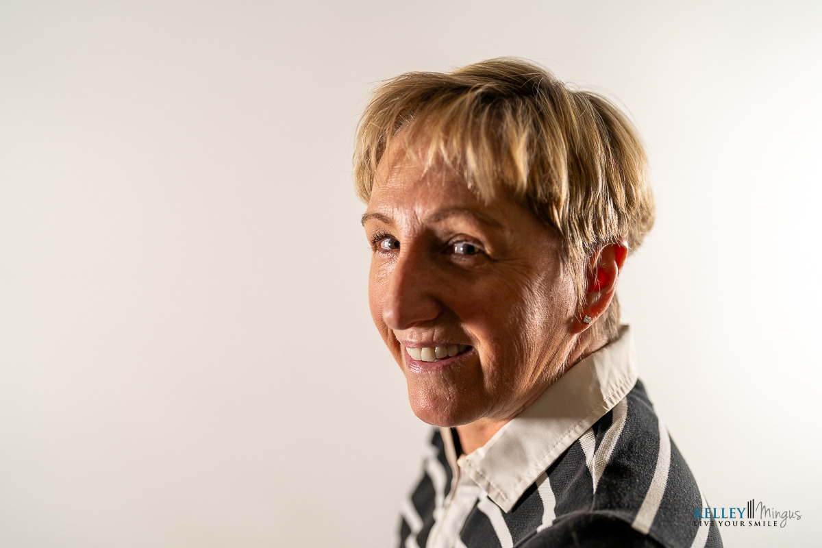 A woman with short blonde hair smiles at the camera, showcasing her radiant results from holistic full mouth rehabilitation, while wearing a black and white striped jacket against a plain white background.
