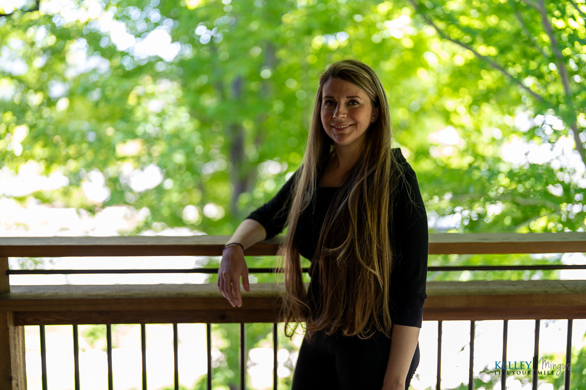 A woman with long hair stands by a wooden railing outdoors, with green trees and sunlight in the background, reflecting on holistic approaches to dental care.