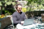 A man sits at an outdoor table using a laptop, smiling while working. The background includes greenery and sunlight, suggesting he’s enjoying a Porcelain Veneers Consultation in a relaxed setting.