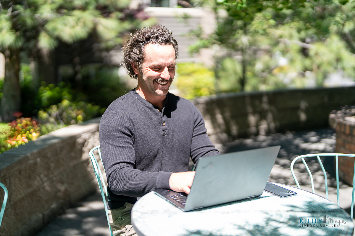 A man sits at an outdoor table using a laptop, smiling while working. The background includes greenery and sunlight, suggesting he’s enjoying a Porcelain Veneers Consultation in a relaxed setting.