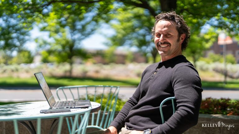 Man sits outdoors at a table with a laptop, smiling at the camera. Surrounded by trees and greenery, he enjoys the sunlight—perhaps taking a break to focus on holistic TMJ relief in this peaceful setting.