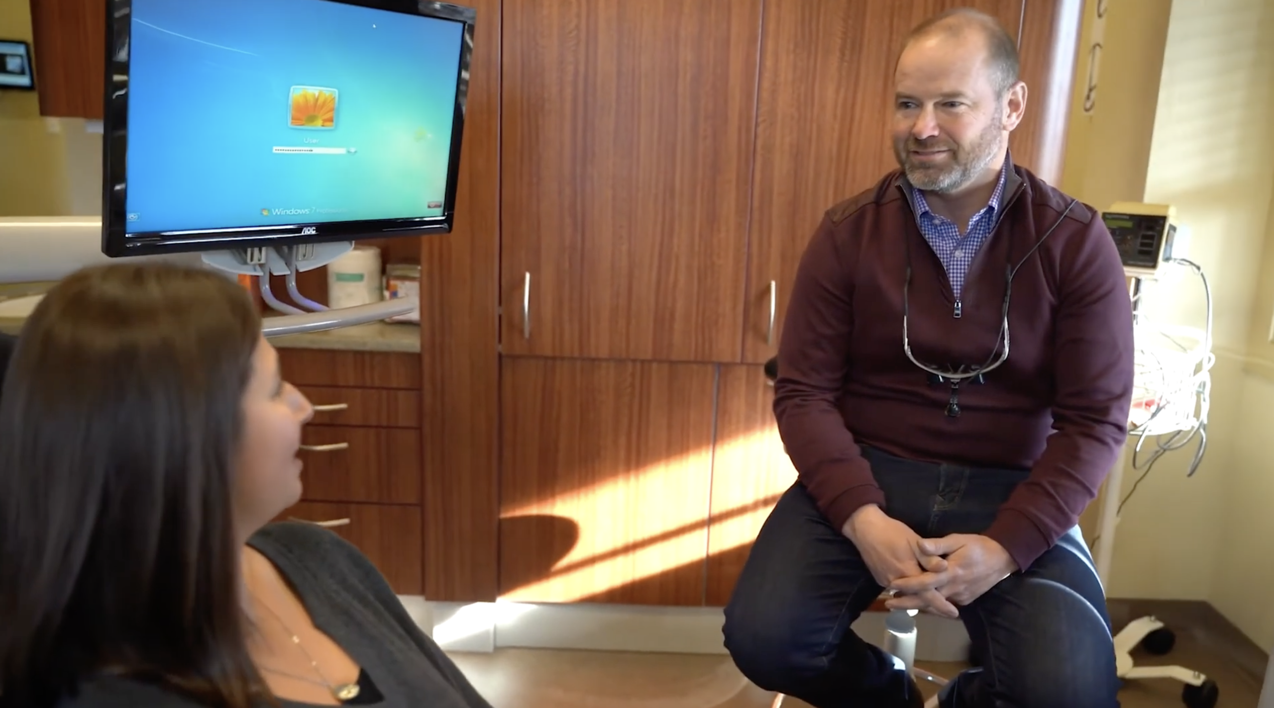 A man in a maroon sweater talks to a seated woman about TMJ relief in a dental office; a computer with a login screen is visible in the background.