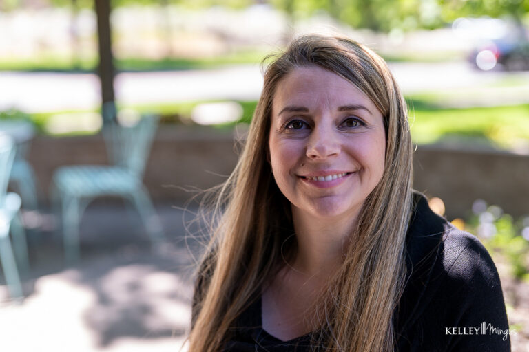 A woman with long blonde hair and a black top sits outdoors, smiling at the camera, perhaps reflecting on her confident smile thanks to metal-free dental restoration options. Empty green metal chairs and blurred greenery are in the background.