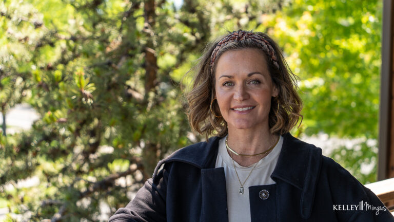 A woman with shoulder-length hair wearing a headband, black coat, and layered necklaces stands outdoors in front of green trees, smiling at the camera—radiating confidence that comes from embracing holistic approaches to dental care.