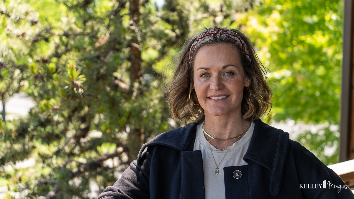 A woman with shoulder-length hair wearing a headband, black coat, and layered necklaces stands outdoors in front of green trees, smiling at the camera—radiating confidence that comes from embracing holistic approaches to dental care.