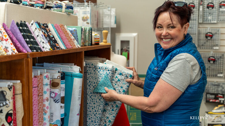 A woman in a blue vest smiles while selecting patterned fabric from a shelf, her confidence shining much like those who benefit from holistic approaches to cosmetic dentistry.