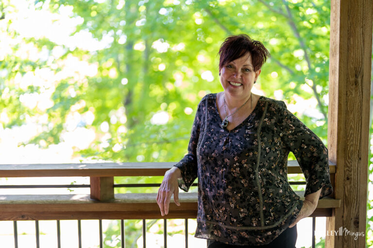 A woman with short dark hair stands smiling on a wooden porch, wearing a dark floral blouse; she radiates calm and confidence, reflecting the benefits of holistic sleep apnea treatment, with green trees blurred in the background.