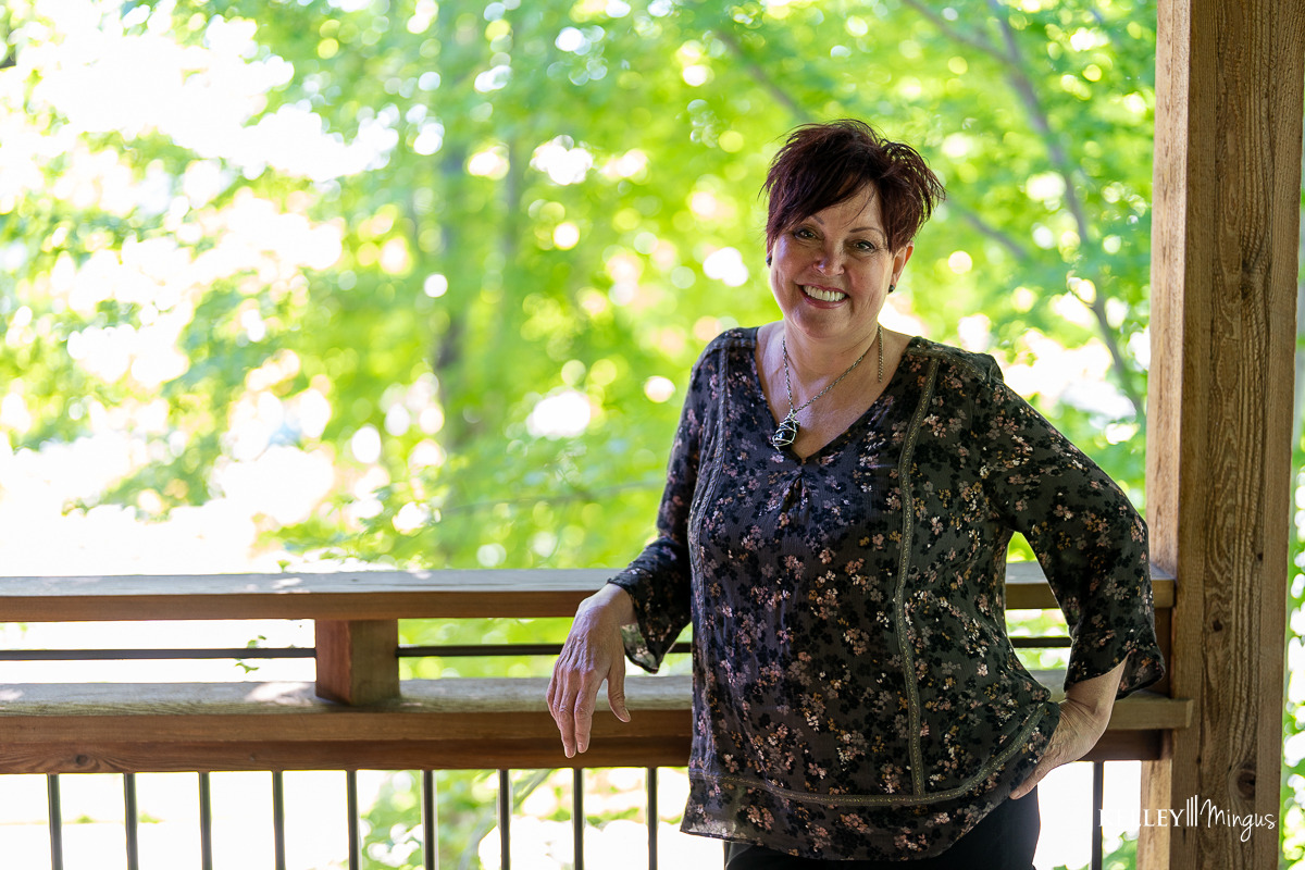 A woman with short dark hair stands smiling on a wooden porch, wearing a dark floral blouse; she radiates calm and confidence, reflecting the benefits of holistic sleep apnea treatment, with green trees blurred in the background.