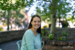 A woman with long brown hair, wearing a light blue top, sits smiling on a metal chair outdoors with trees and a brick planter in the background, looking as cheerful as if she just visited her family dentist in Bend Oregon.