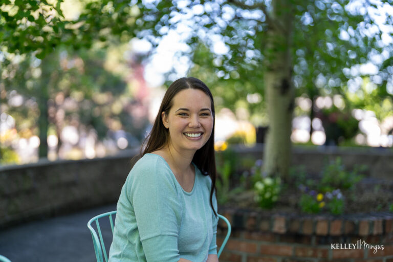 A woman with long brown hair, wearing a light blue top, sits smiling on a metal chair outdoors with trees and a brick planter in the background, looking as cheerful as if she just visited her family dentist in Bend Oregon.