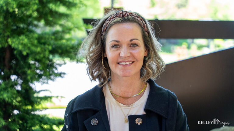 A woman with shoulder-length wavy hair wearing a headband, light shirt, dark jacket, and necklaces stands outdoors, smiling at the camera after experiencing relief from TMJ treatment for migraines.