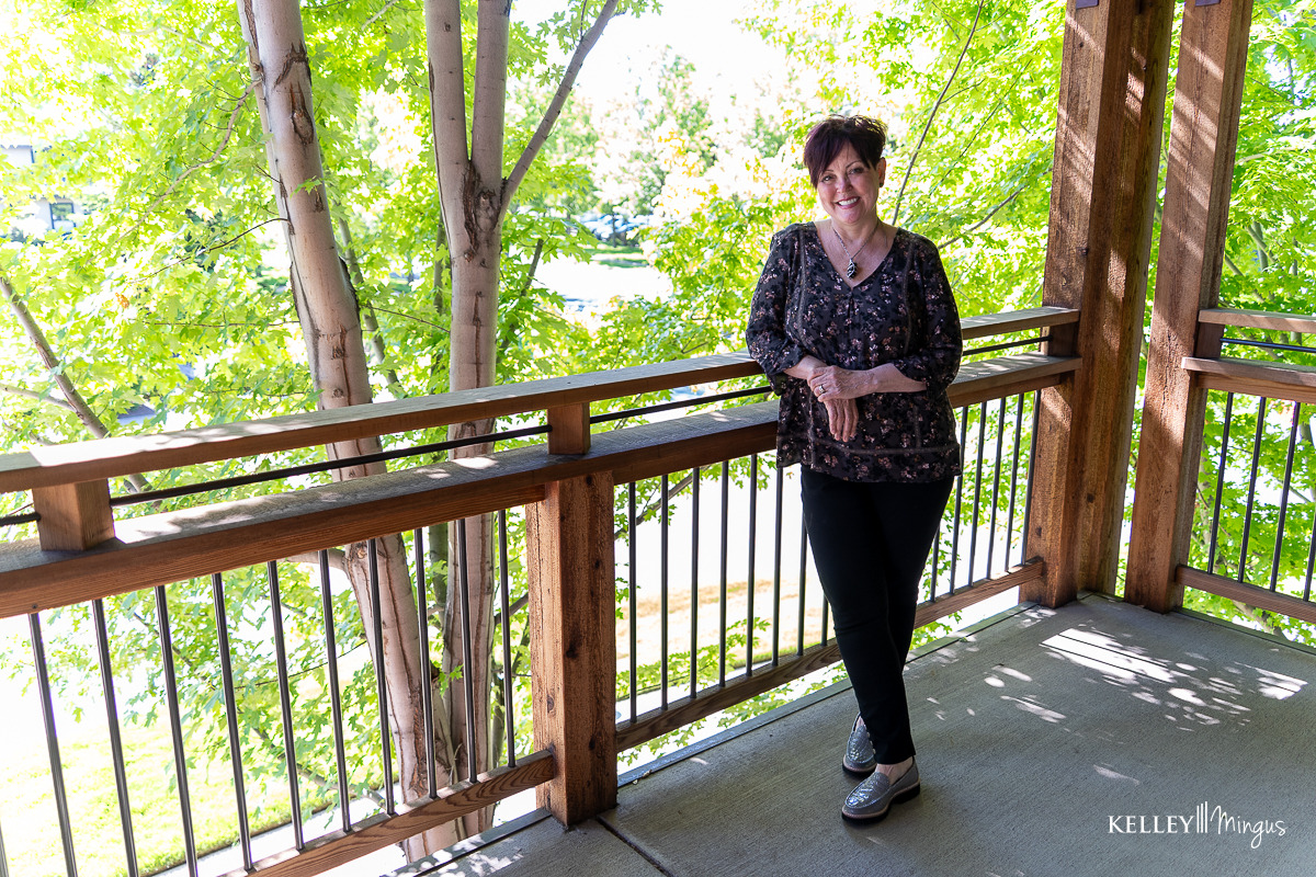 A woman stands on a wooden balcony surrounded by green trees, smiling at the camera in daylight—radiating confidence thanks to holistic approaches to cosmetic dentistry.