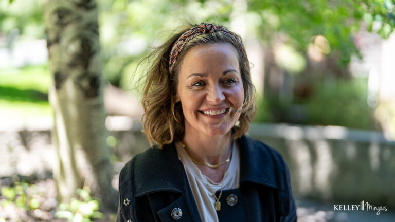 A woman with shoulder-length hair and a headband stands outdoors, smiling at the camera. She is wearing a dark jacket and layered necklaces, enjoying fresh air after TMJ treatment for migraines. Blurred greenery is in the background.