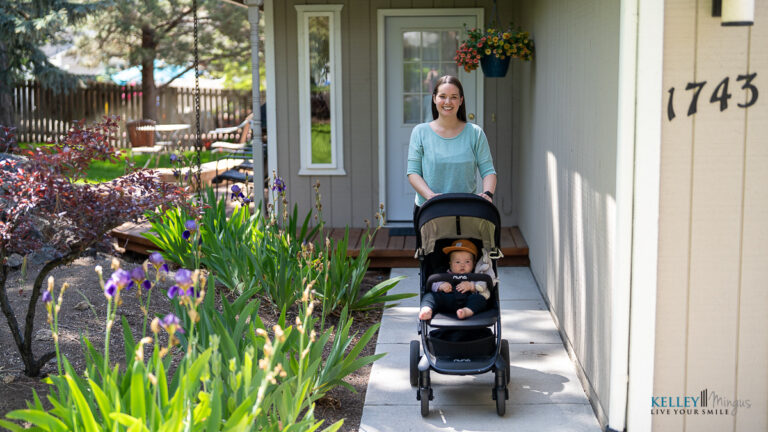 A woman pushes a baby in a stroller along a garden path outside a house with the number 1743 visible on the wall, reminiscent of exploring metal-free dental restoration options for a healthier future.
