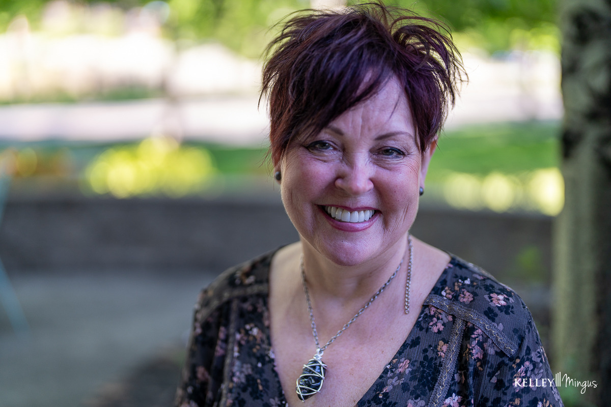 A woman with short, dark reddish hair smiles outdoors, wearing a patterned top and a pendant necklace; greenery is visible in the blurred background, evoking the calm often sought in holistic sleep apnea treatment.
