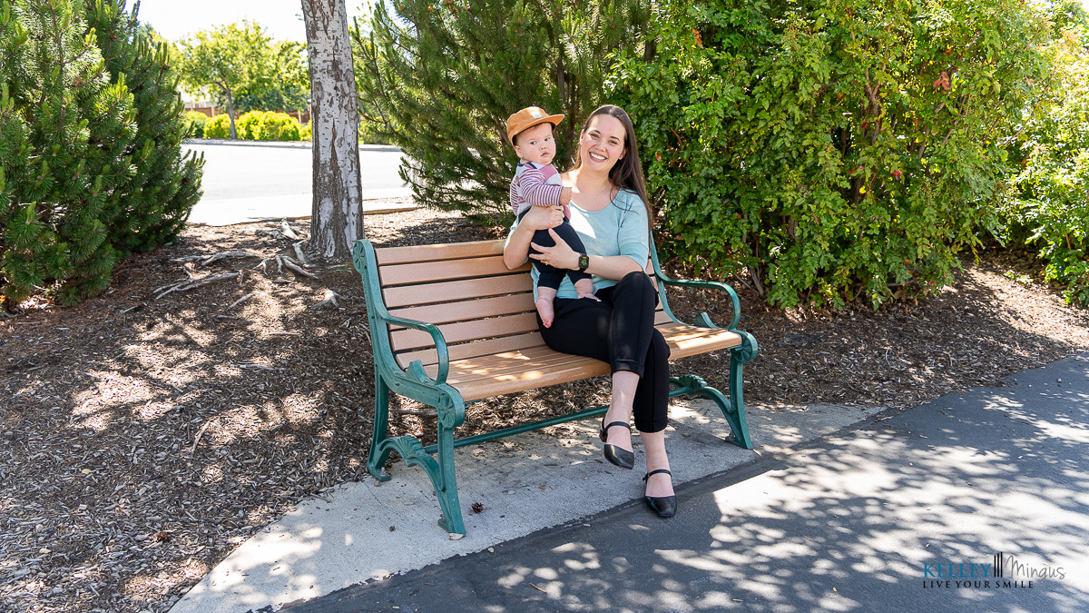 A woman sits on a park bench holding a baby on her lap. Both are smiling and surrounded by trees and greenery, enjoying the moment while discussing metal-free dental restoration options for a healthier lifestyle.