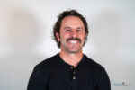 A man with curly hair and a mustache is smiling at the camera, wearing a black henley shirt against a plain light background, looking confident after successful TMJ treatment for migraines.
