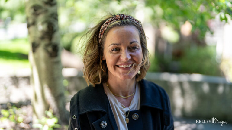 A woman with wavy hair and a headband smiles outdoors, enjoying relief from TMJ treatment for migraines. She wears a dark coat and layered necklaces, with blurred trees and greenery in the background.