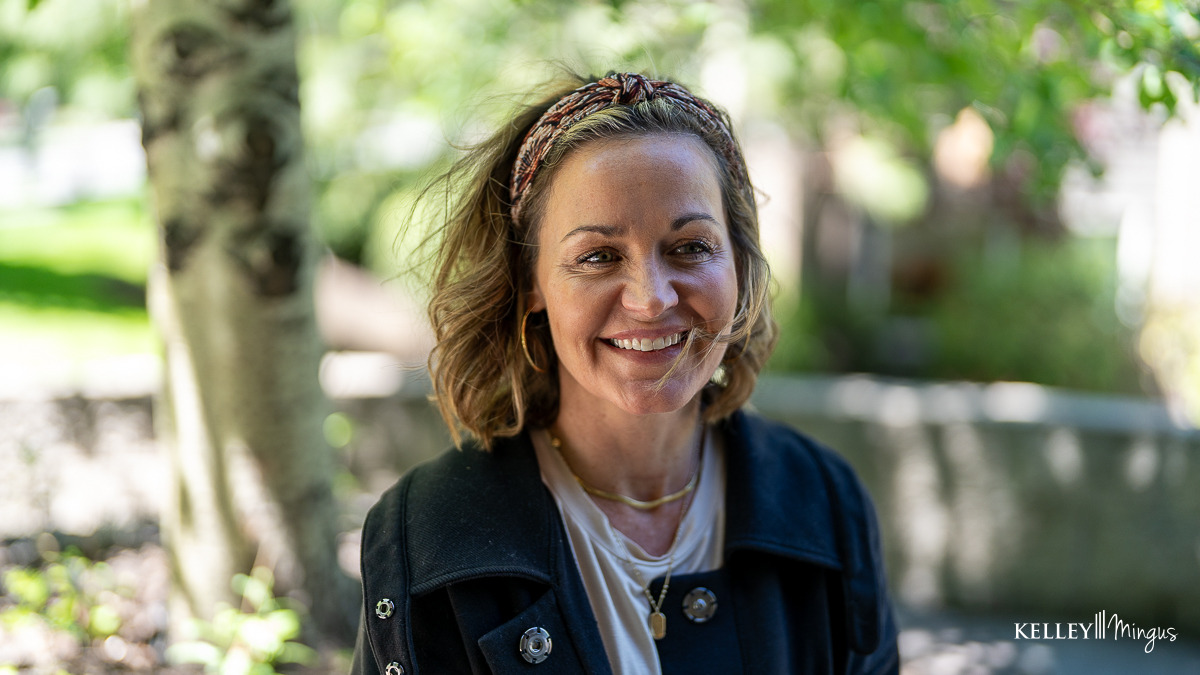 A woman with wavy hair and a headband smiles outdoors, enjoying relief from TMJ treatment for migraines. She wears a dark coat and layered necklaces, with blurred trees and greenery in the background.