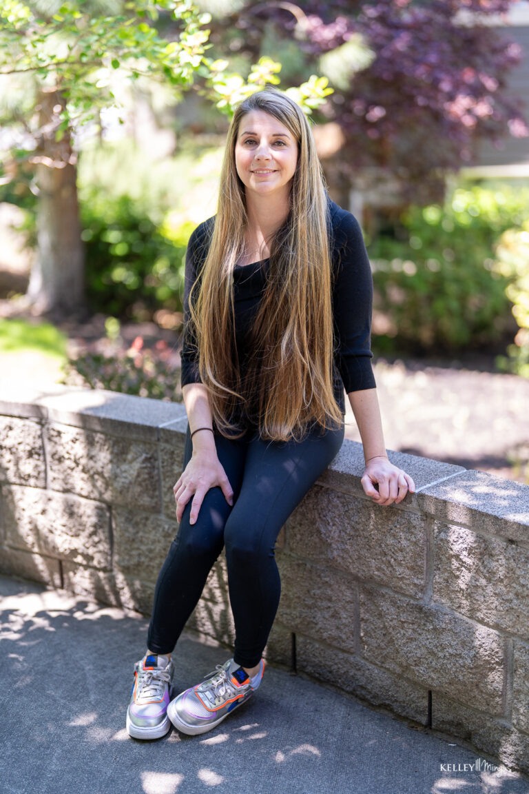 A woman with long straight hair, wearing a black top and leggings with silver sneakers, sits on a low stone wall outdoors in daylight, perhaps reflecting on holistic approaches to dental care.