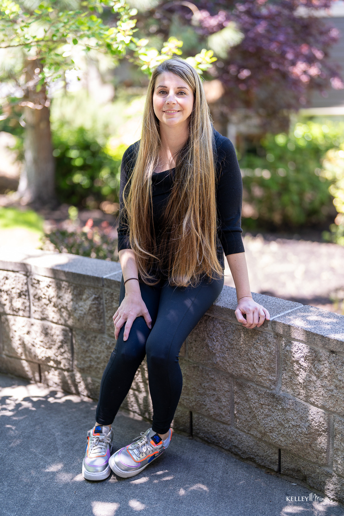 A woman with long straight hair, wearing a black top and leggings with silver sneakers, sits on a low stone wall outdoors in daylight, perhaps reflecting on holistic approaches to dental care.