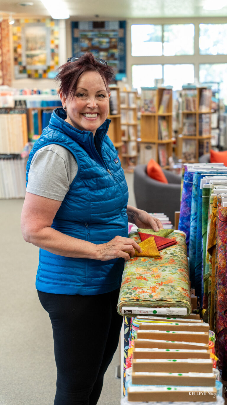 Woman smiling while arranging colorful fabric bolts in a well-lit fabric store, her radiant smile as bright as one from a Bend Oregon cosmetic dentist, with shelves and quilts visible in the background.