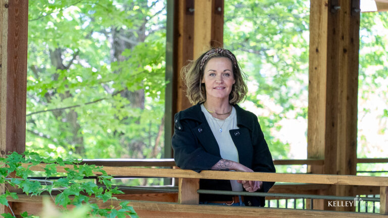A woman with shoulder-length hair stands and smiles by a wooden railing in an outdoor pavilion surrounded by green trees, radiating the natural confidence often associated with holistic dentistry.