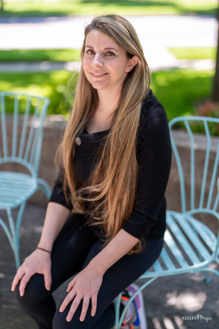 A woman with long blonde hair sits on a light blue metal chair outdoors in Bend, Oregon, wearing a black top and pants, with a blurred green background.