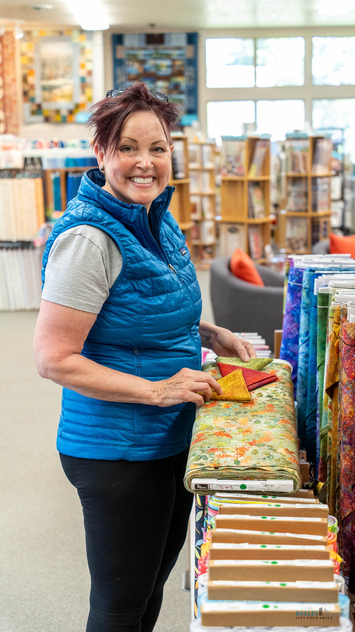 A woman wearing a blue vest stands in a fabric store, holding a piece of yellow cloth over a roll of floral fabric and smiling as if sharing tips on how long do porcelain veneers last.