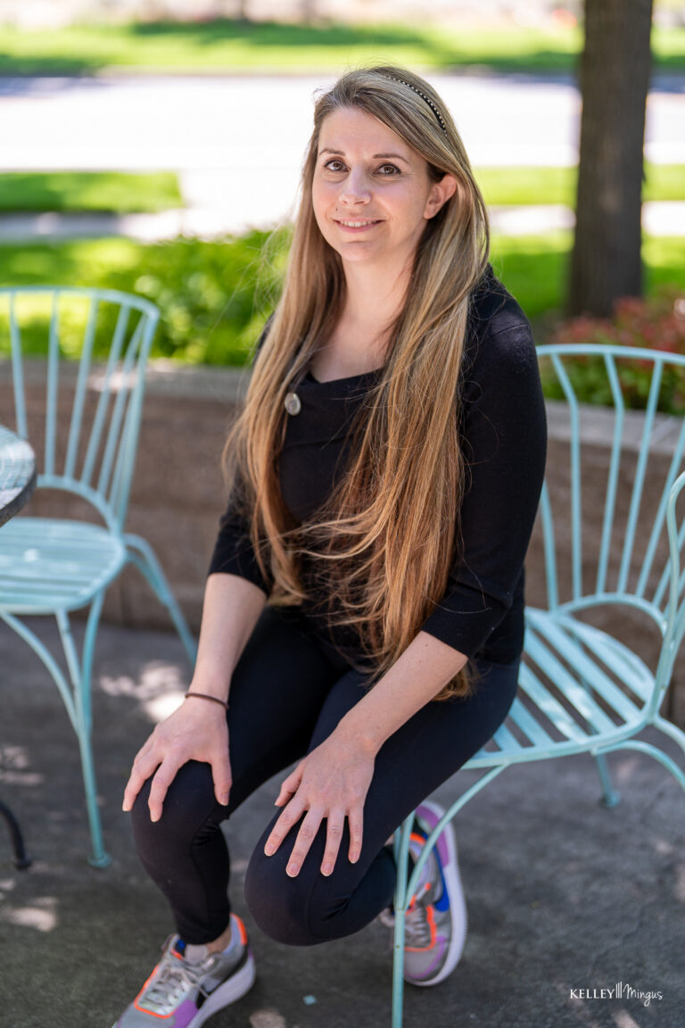 A woman with long blonde hair, wearing a black top and leggings, sits on a light blue metal chair outdoors near a table, greenery in the background—relaxed after her visit to a family dentist in Bend Oregon.