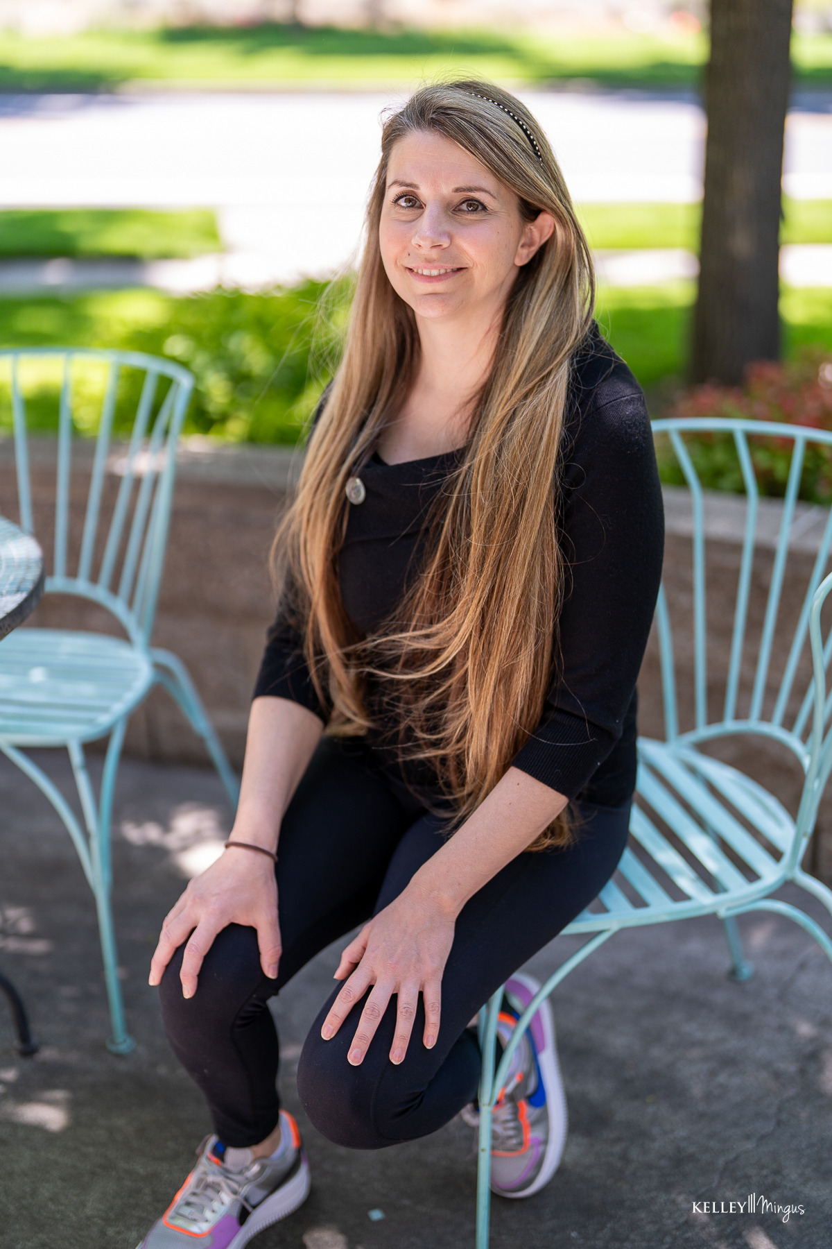 A woman with long blonde hair, wearing a black top and leggings, sits on a light blue metal chair outdoors near a table, greenery in the background—relaxed after her visit to a family dentist in Bend Oregon.