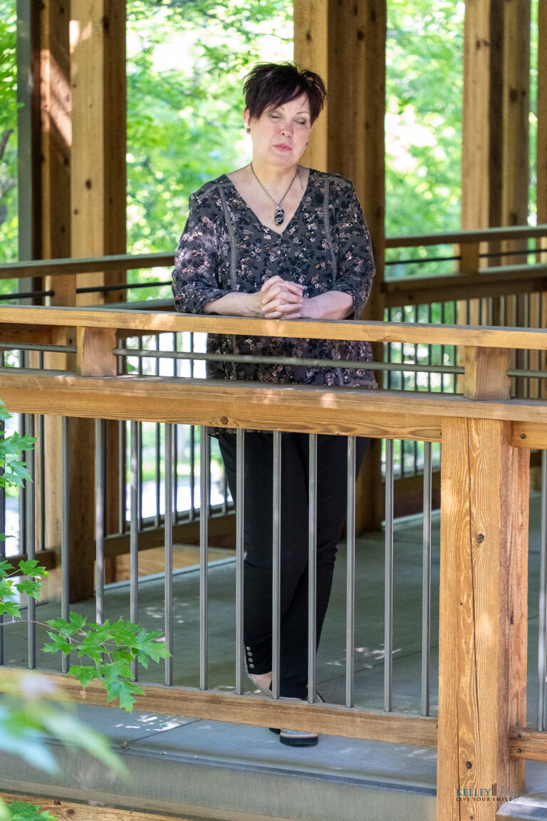 A woman with short dark hair stands on a wooden walkway, leaning on a railing, surrounded by green trees, lost in thought about full mouth rehabilitation vs. smile makeover.