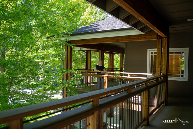 A person stands on the wooden porch of a house surrounded by green trees on a sunny day, enjoying the peaceful atmosphere after a visit to a holistic dentistry clinic.