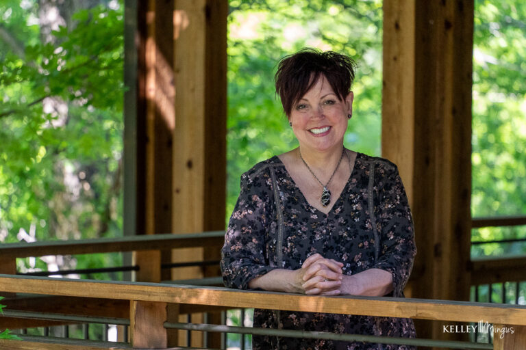 A woman with short dark hair and a floral blouse stands smiling, leaning on a wooden railing in a shaded, outdoor setting with green foliage—perhaps awaiting an appointment with Kelley Mingus, sleep apnea specialist.