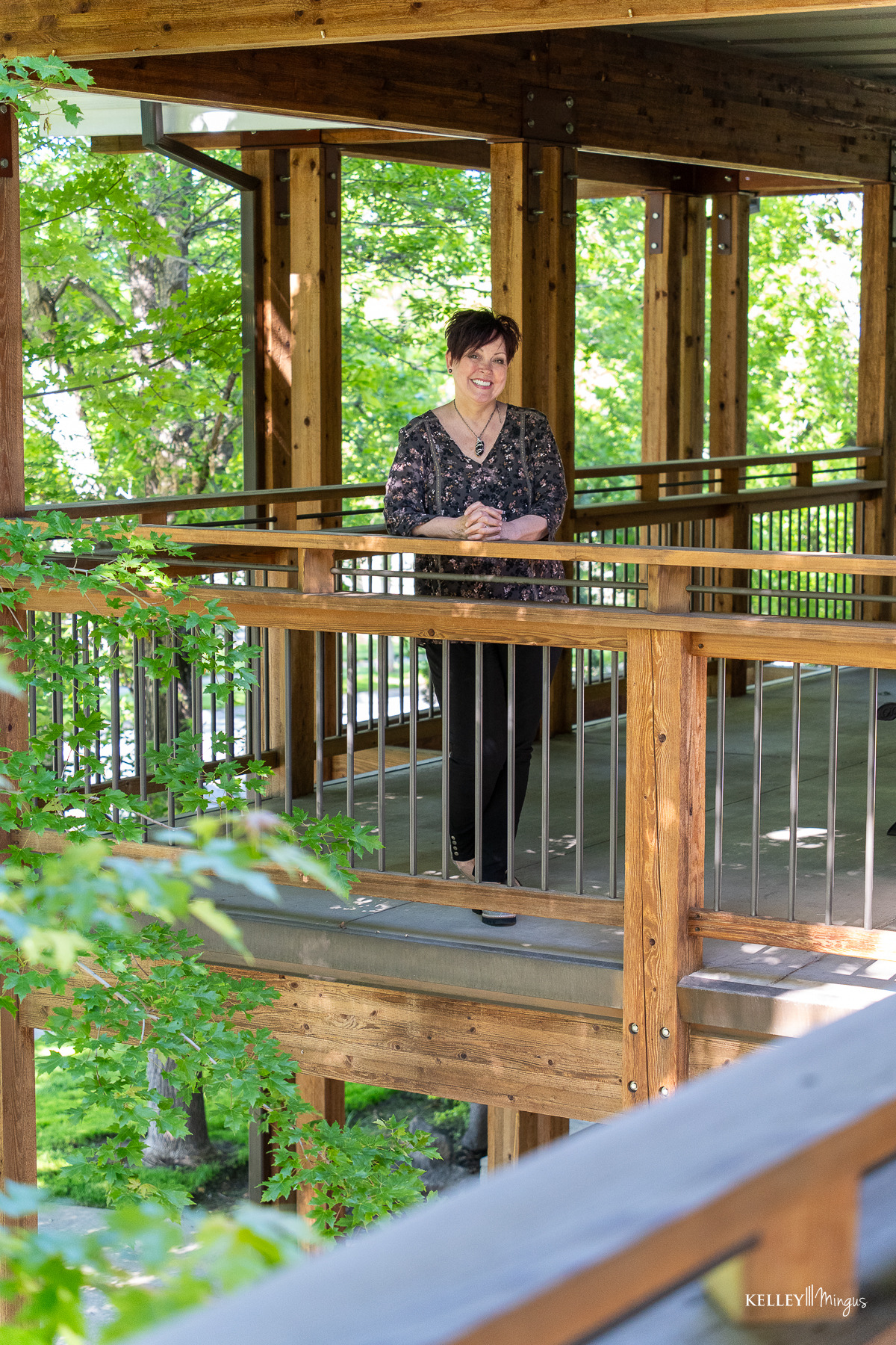 A woman stands on a wooden walkway surrounded by green trees, smiling and looking into the distance—perhaps imagining her next visit to the best TMJ specialist in Bend.