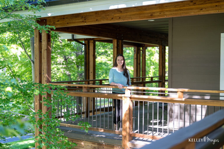 A woman stands and smiles on a wooden porch surrounded by green trees on a sunny day, enjoying the confidence that comes from visiting her trusted family dentist in Bend, Oregon.