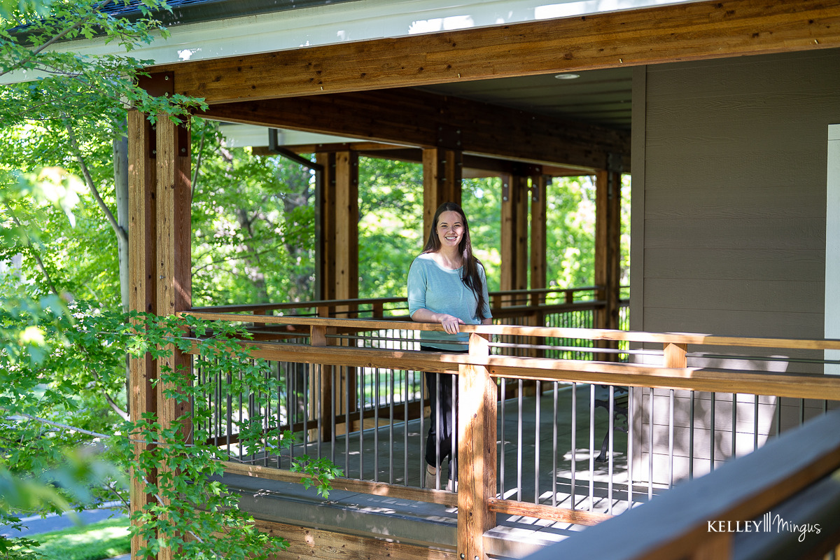 A woman stands and smiles on a wooden porch surrounded by green trees on a sunny day, enjoying the confidence that comes from visiting her trusted family dentist in Bend, Oregon.