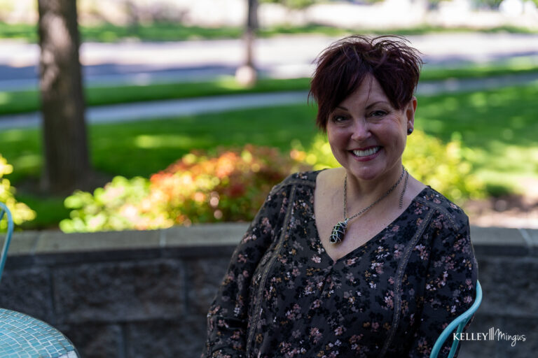 A woman with short dark hair sits outdoors on a patio, smiling at the camera—a smile that could be achieved through a custom smile design process. Green grass and trees are visible in the background.