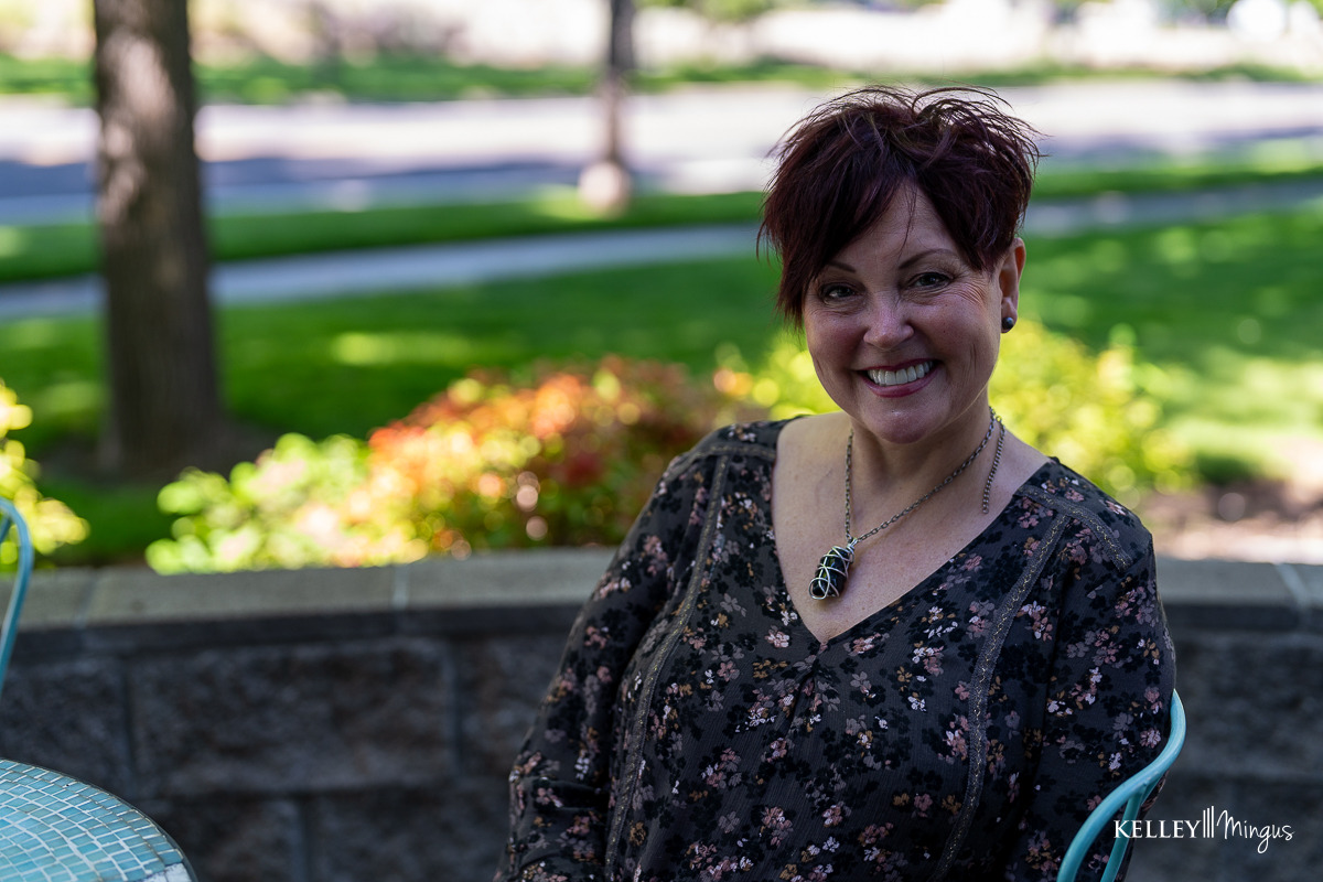 A woman with short dark hair sits outdoors on a patio, smiling at the camera—a smile that could be achieved through a custom smile design process. Green grass and trees are visible in the background.