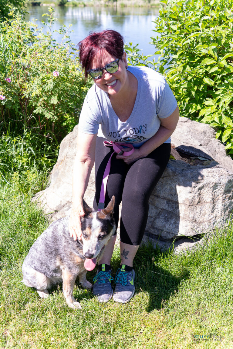 A woman wearing sunglasses and a "Not Today" t-shirt sits on a rock near water, smiling and petting her gray and brown dog. The Bend Oregon cosmetic dentist enjoys a sunny day in the grassy area with her furry friend.