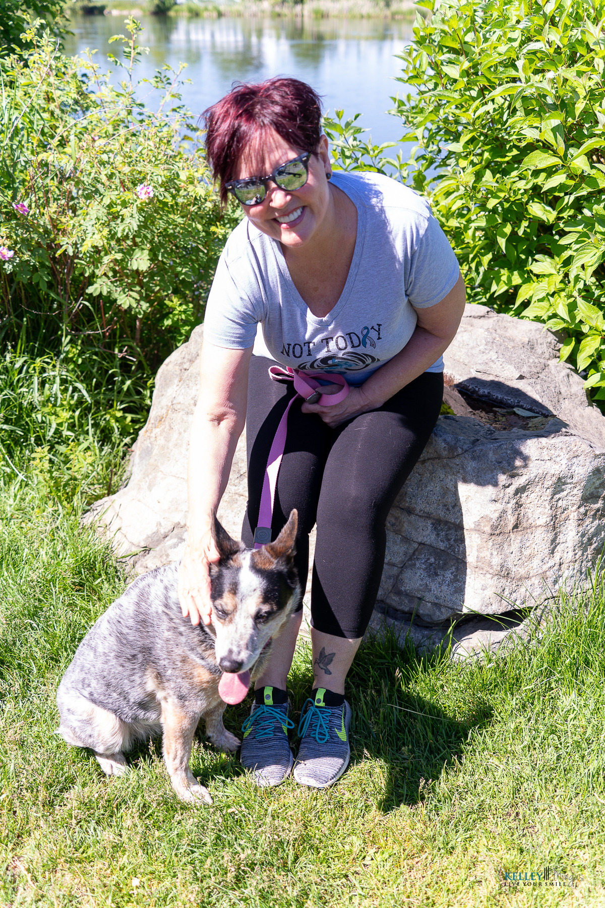 A woman wearing sunglasses and a "Not Today" t-shirt sits on a rock near water, smiling and petting her gray and brown dog. The Bend Oregon cosmetic dentist enjoys a sunny day in the grassy area with her furry friend.