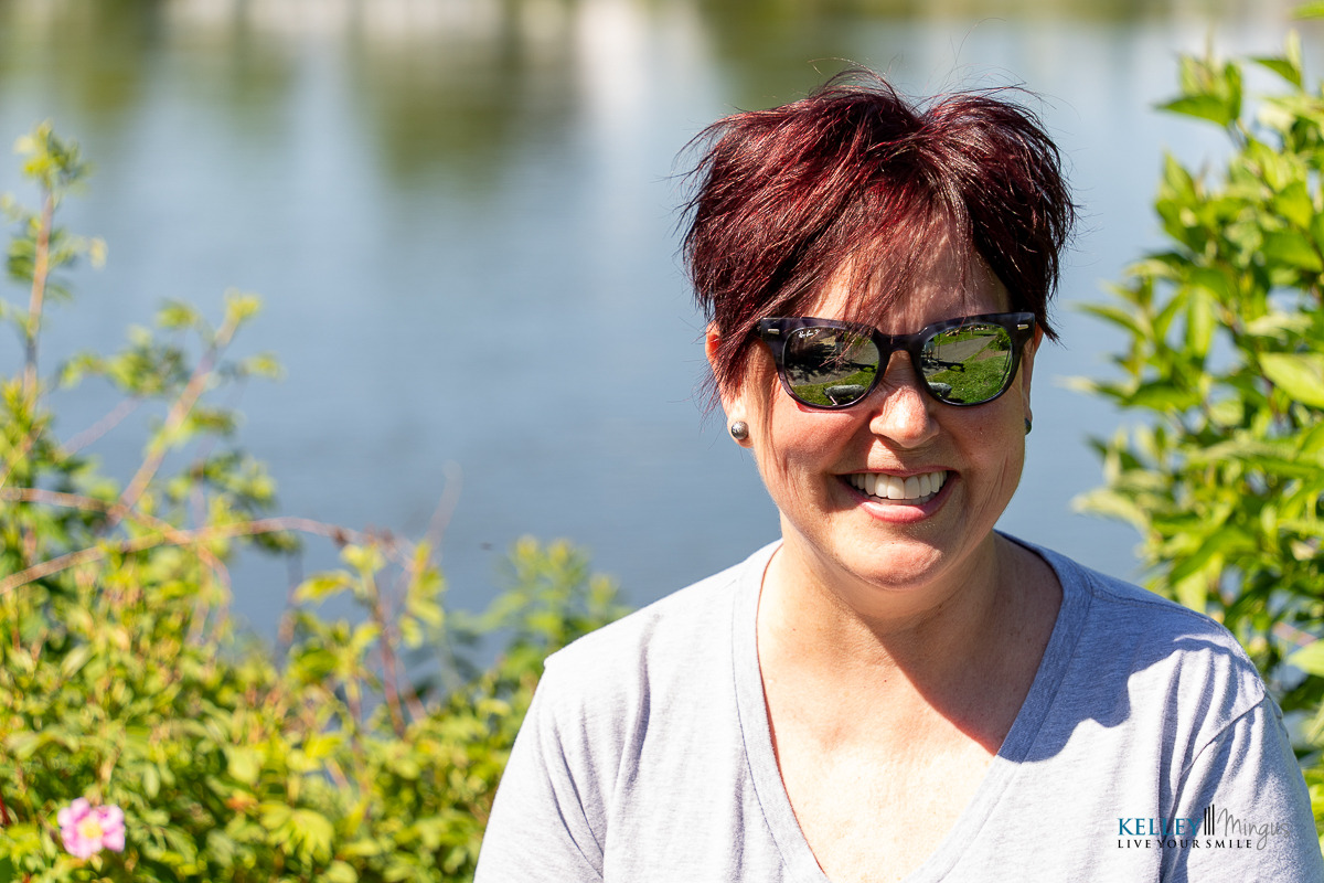 A woman with short red hair and sunglasses, flashing a radiant smile outdoors in front of greenery and a body of water, showcases the confidence inspired by her Bend Oregon cosmetic dentist on a sunny day.