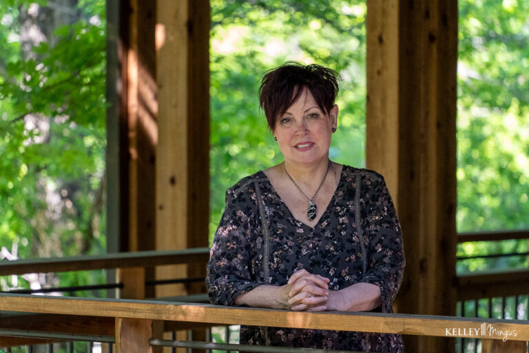 A woman with short dark hair, wearing a patterned blouse and necklace, stands indoors by a wooden railing with greenery outside—perhaps contemplating full mouth rehabilitation vs. smile makeover options.