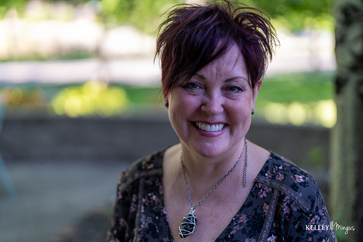 A woman with short reddish hair and a floral top smiles outdoors, her pendant necklace catching the light. The blurred greenery in the background adds to her vibrant look—much like how long do porcelain veneers last, enhancing a smile for years.