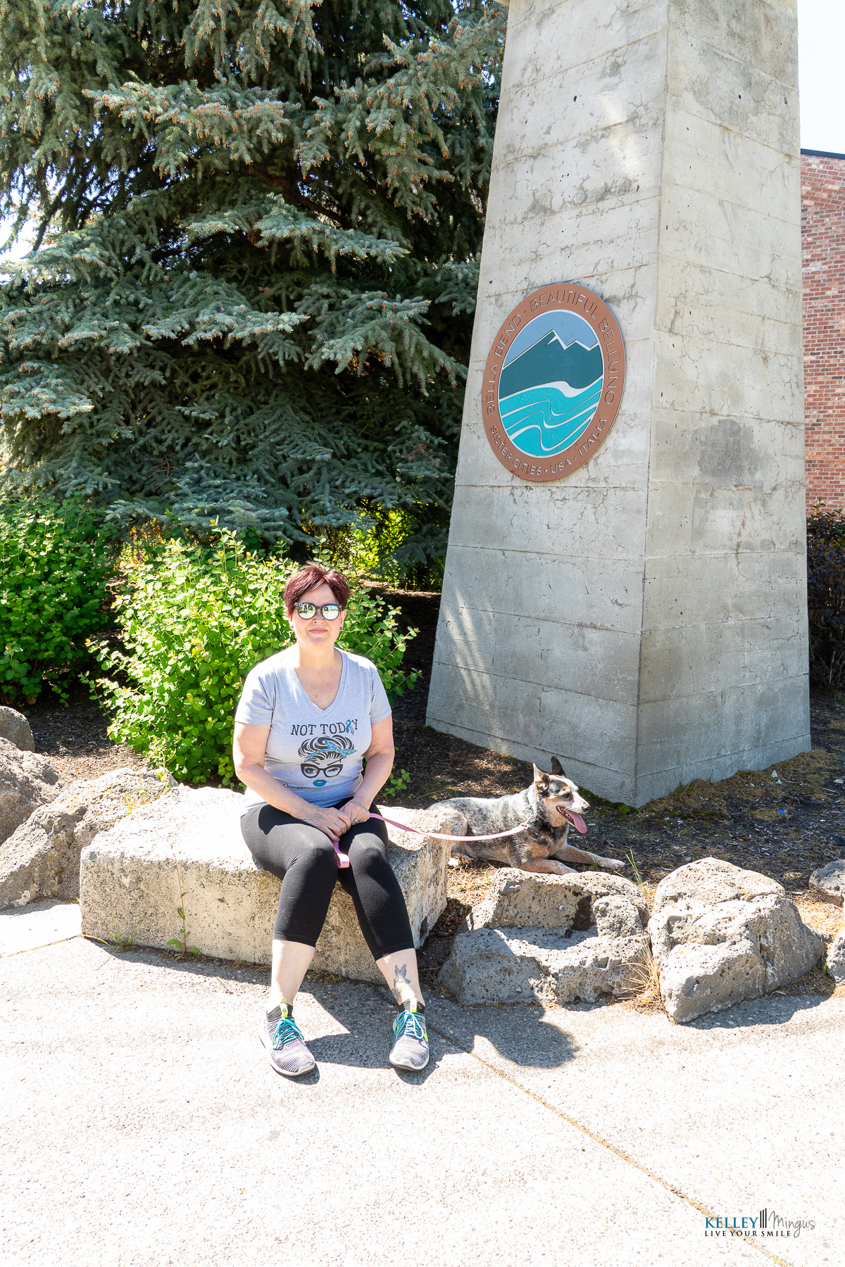 A person wearing sunglasses and a gray T-shirt sits on a stone ledge next to a dog, near a concrete monument with a circular emblem outdoors, perhaps reflecting after visiting Kelley Mingus, sleep apnea specialist.