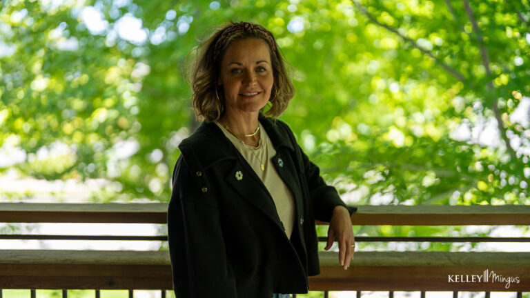 A woman in a black jacket stands by a wooden railing outdoors, with green leafy trees in the background, perhaps reflecting on the benefits of holistic dentistry and natural wellness.